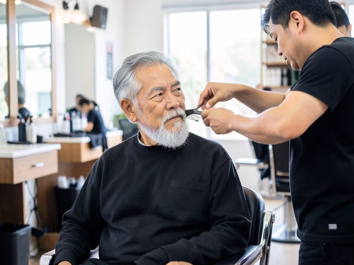 Barber trimming an older man's gray beard in a bright barbershop